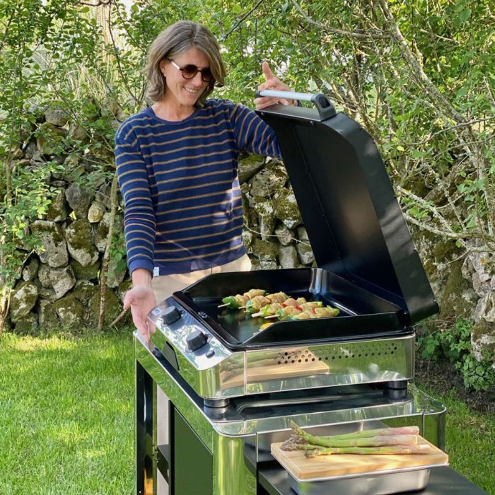 Femme ouvrant un couvercle de protection noir sur une plancha inox en extérieur.