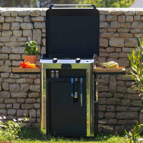 Plancha Gaz Fusion 60 sur chariot inox en extérieur, avec tablettes rabattables et accessoires de cuisine.