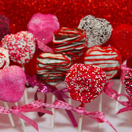 Assortiment de cake pops en forme de cœurs décorés de glaçage rouge, blanc et rose, avec des paillettes et des vermicelles colorés, présentés sur des bâtonnets ornés de rubans roses, parfaits pour la Saint-Valentin.