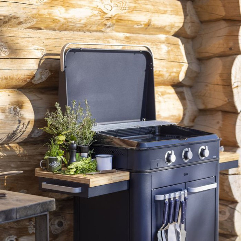 Plancha ENOSIGN 80 bleu navy - ENO avec couvercle ouvert, installée sur un meuble extérieur, espace de préparation avec planche en bois et herbes aromatiques.