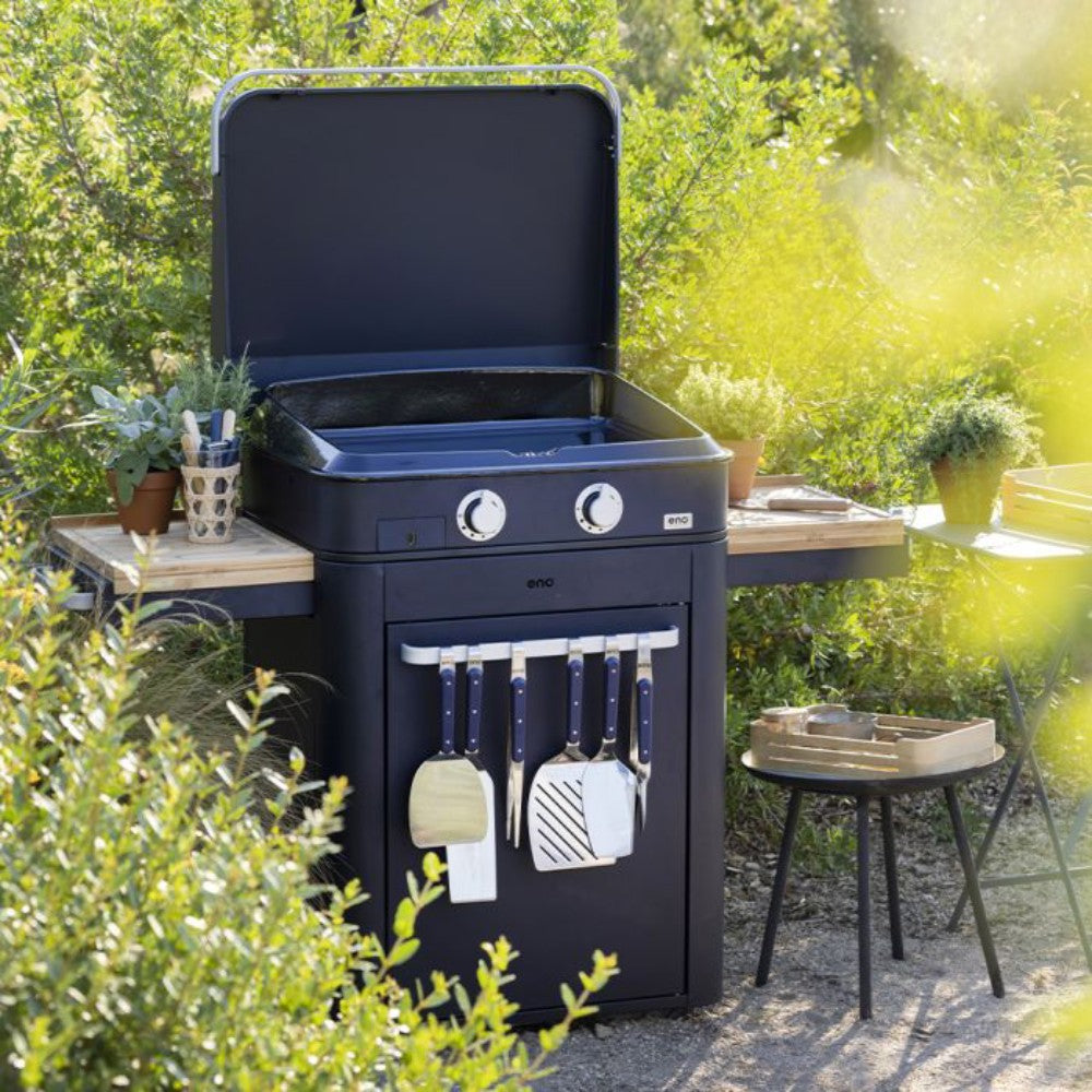 Plancha Enosign 65 bleu navy installée en extérieur, équipée d’ustensiles et de tablettes latérales en bois, parfaite pour des repas en plein air.